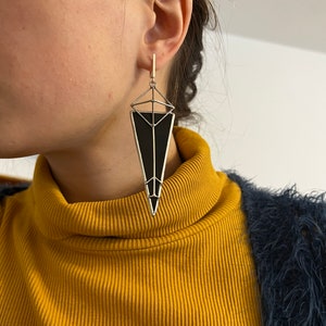 May include: Geometric black and silver earrings. The earrings have a long, triangular shape with a black center and silver metal frame. The earrings are hanging from a gold-colored ear wire. The design is modern and stylish.