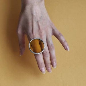 May include: Three orange slices on a yellow background. The orange slices are arranged in a triangular shape. A hand with a large silver ring is visible in the top left corner of the image.
