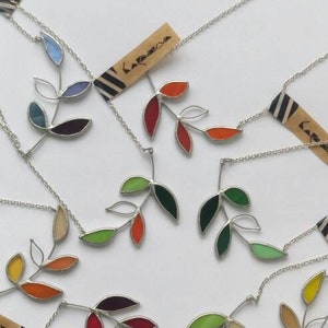 Puede incluir: Una colección de diez collares de plata con colgantes de hojas de vidrio de colores. Cada collar tiene una pequeña etiqueta con la palabra "Lafeuca" escrita en ella.