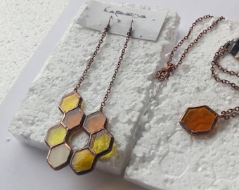 Puede incluir: Dos collares con colgantes de vidrio en forma de panal. Un collar tiene un solo colgante grande de ámbar, el otro tiene un grupo de colgantes más pequeños de color amarillo y ámbar. Ambos collares están hechos con cadena de cobre.