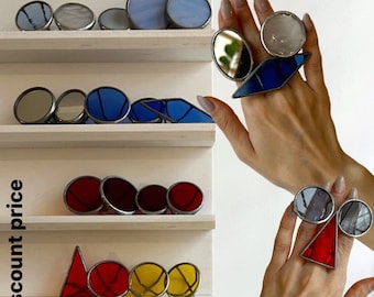 May include: Assortment of colorful stained glass rings on display. The rings feature various shapes and designs, including circles, triangles, and geometric patterns. Available rings with discount price.