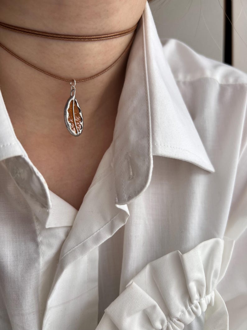 May include: A brown leather choker necklace with a silver pendant shaped like a leaf. The leaf is filled with a translucent orange material.