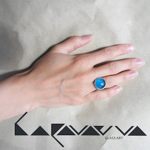 May include: A silver ring with a round, light blue glass stone. The ring is on a person's finger. The background is a light brown surface. The logo "LAVAVA GLASS ART" is in the lower left corner.