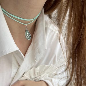 May include: A delicate silver necklace with a light green leaf pendant. The necklace is layered on a light blue cord.