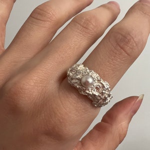 Könnte beinhalten: Ein silberner Ring mit einem strukturierten, organischen Design. Der Ring hat eine kleine, runde, hellfarbene Perle in der Mitte. Der Ring hat eine raue, unebene Oberfläche, die ihm ein einzigartiges, handgefertigtes Aussehen verleiht. Schmuck.