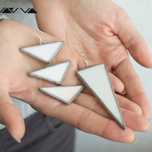 May include: A pair of white geometric earrings with silver accents. The earrings are made of glass and are shaped like triangles. The earrings are hanging from a person's hand.