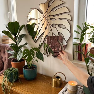 Pode incluir: Um espelho decorativo em forma de folha grande, com uma moldura de metal castanho, est&aacute; pendurado numa parede branca. O espelho reflete a divis&atilde;o e &eacute; rodeado por plantas em vasos, um regador dourado e uma vela acesa. Um elemento decorativo.