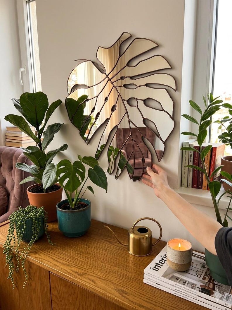 Pode incluir: Um espelho decorativo em forma de folha grande, com uma moldura de metal castanho, est&aacute; pendurado numa parede branca. O espelho reflete a divis&atilde;o e &eacute; rodeado por plantas em vasos, um regador dourado e uma vela acesa. Um elemento decorativo.