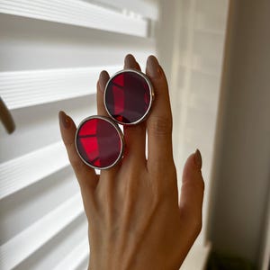 May include: Two large, round, red gemstone rings with silver-colored metal settings. The rings are displayed on a hand, showcasing their size and color. The gemstones have a faceted appearance, catching the light.