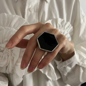 May include: A large, geometric ring with a black, hexagonal center stone and a silver-colored metal frame. The ring is worn on a finger, set against a white ruffled shirt.