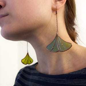 Puede incluir: Pendientes de color cobre con diseño de hoja de ginkgo. Los pendientes presentan una cadena y una hoja en forma de abanico con una combinación de colores verde y amarillo. La persona lleva una camiseta a rayas azules y blancas.