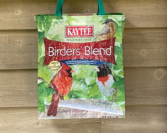 May include: A green tote bag made from recycled birdseed packaging. The bag features a picture of a cardinal, a grosbeak, and a chickadee, along with the text "Kaytee Wild Bird Food Birders' Blend".