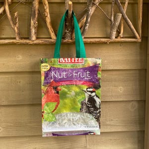 May include: A green tote bag made from recycled birdseed packaging. The bag features a colorful image of a cardinal and a woodpecker with the text "Mille Wild Bird Food Nut & Fruit Blend" printed on the front.