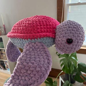 May include: A crocheted stuffed turtle toy with a pink, blue, and purple color scheme. The turtle has a large, round shell and a small head with big eyes.