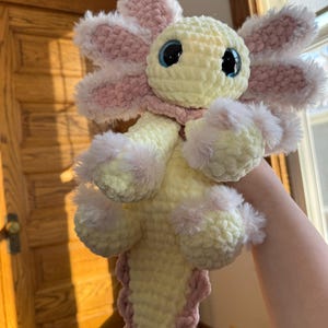 Puede incluir: Un juguete de axolotl de peluche hecho a mano en amarillo suave, rosa y blanco. La criatura de ganchillo presenta grandes ojos azules, branquias esponjosas y una cola con volantes. El axolotl es un juguete caprichoso y tierno.