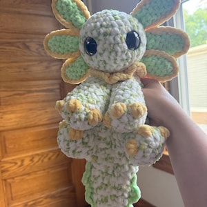 May include: A handmade crochet axolotl plushie in shades of white, green, and yellow. The plushie features large black eyes and a frilly, flower-like head. The axolotl's body is detailed with textured stitches.
