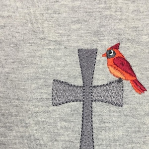 May include: Embroidered design of a cardinal perched on a gray cross. The cardinal is red with a black eye and beak. The cross is a simple, stylized design.