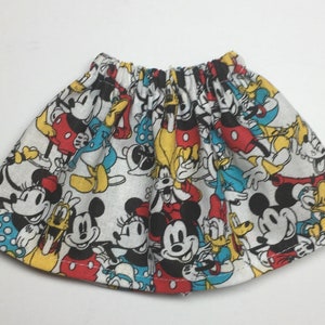 May include: A white, elastic-waist skirt featuring a colorful pattern of Mickey Mouse, Minnie Mouse, Donald Duck, and other Disney characters. The skirt has a gathered waist and a flared shape, with red, yellow, black, and blue accents.