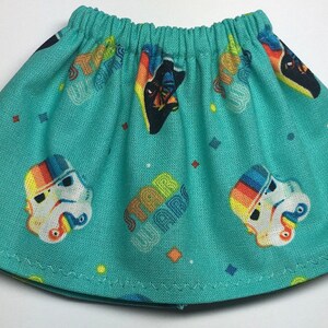 May include: A turquoise skirt with an elastic waistband, featuring a repeating pattern of Stormtrooper helmets with rainbow accents, the words "STAR WARS", and geometric shapes. The skirt is made of a lightweight fabric.