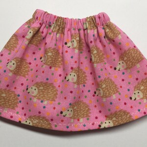 May include: A pink skirt with an elastic waistband, decorated with a repeating pattern of smiling hedgehogs and colorful polka dots. The hem is finished with a white zigzag stitch. This skirt is suitable for a doll.