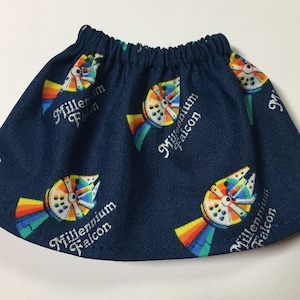 May include: A navy blue skirt with an elastic waistband, patterned with colorful illustrations of the Millennium Falcon and the text "Millennium Falcon" in white. The skirt is a fun, themed apparel item.