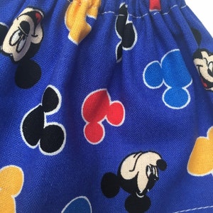 May include: A blue fabric item with a repeating pattern of Mickey Mouse faces, yellow gloves, and Mickey Mouse-shaped designs in red, black, and blue. The fabric is gathered at the top, creating a ruffled effect.