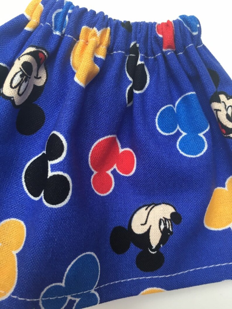 May include: A blue fabric item with a repeating pattern of Mickey Mouse faces, yellow gloves, and Mickey Mouse-shaped designs in red, black, and blue. The fabric is gathered at the top, creating a ruffled effect.