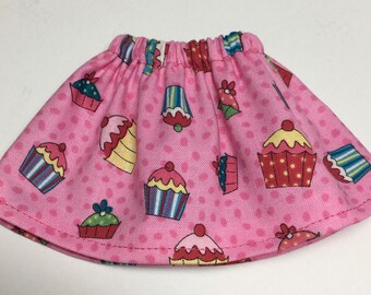 May include: A pink doll skirt with an elastic waistband, covered in a pattern of colorful cupcakes. The cupcakes have various designs and colors, including red, blue, yellow, and green, set against a pink polka dot background.