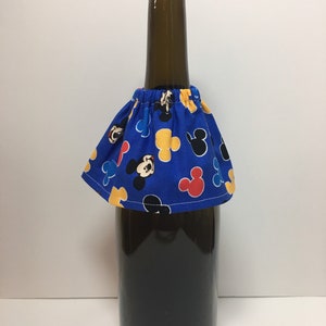 May include: A dark glass bottle with a brown cap is adorned with a blue fabric skirt. The skirt features a pattern of Mickey Mouse in red, yellow, and black. The bottle appears to be filled with a dark liquid.