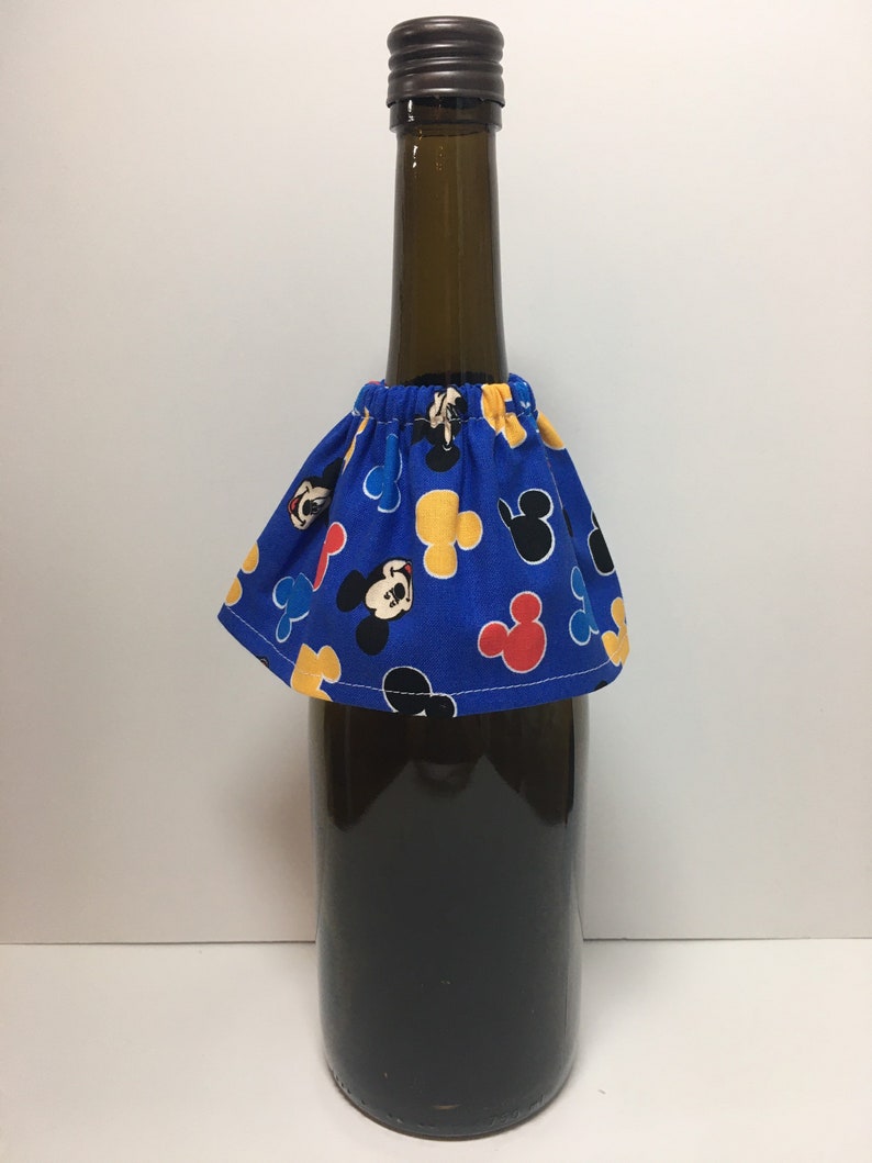 May include: A dark glass bottle with a brown cap is adorned with a blue fabric skirt. The skirt features a pattern of Mickey Mouse in red, yellow, and black. The bottle appears to be filled with a dark liquid.