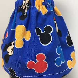 May include: A blue fabric bottle skirt with a repeating pattern of Mickey Mouse heads in black, yellow, red, and blue. The skirt is gathered at the top and has a white stitched hem.