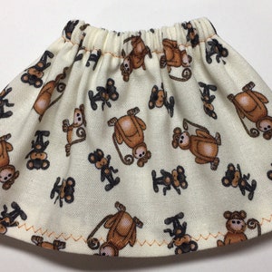 May include: A cream-colored doll skirt with an elastic waistband and a brown monkey pattern. The skirt has a zig-zag stitched hem in orange thread. The monkeys are in various poses.