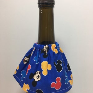 May include: A blue fabric wine bottle skirt featuring a pattern of Mickey Mouse faces and shapes in red, yellow, and black. The skirt is gathered at the top and fits around the neck of a wine bottle. The bottle has a brown cap.