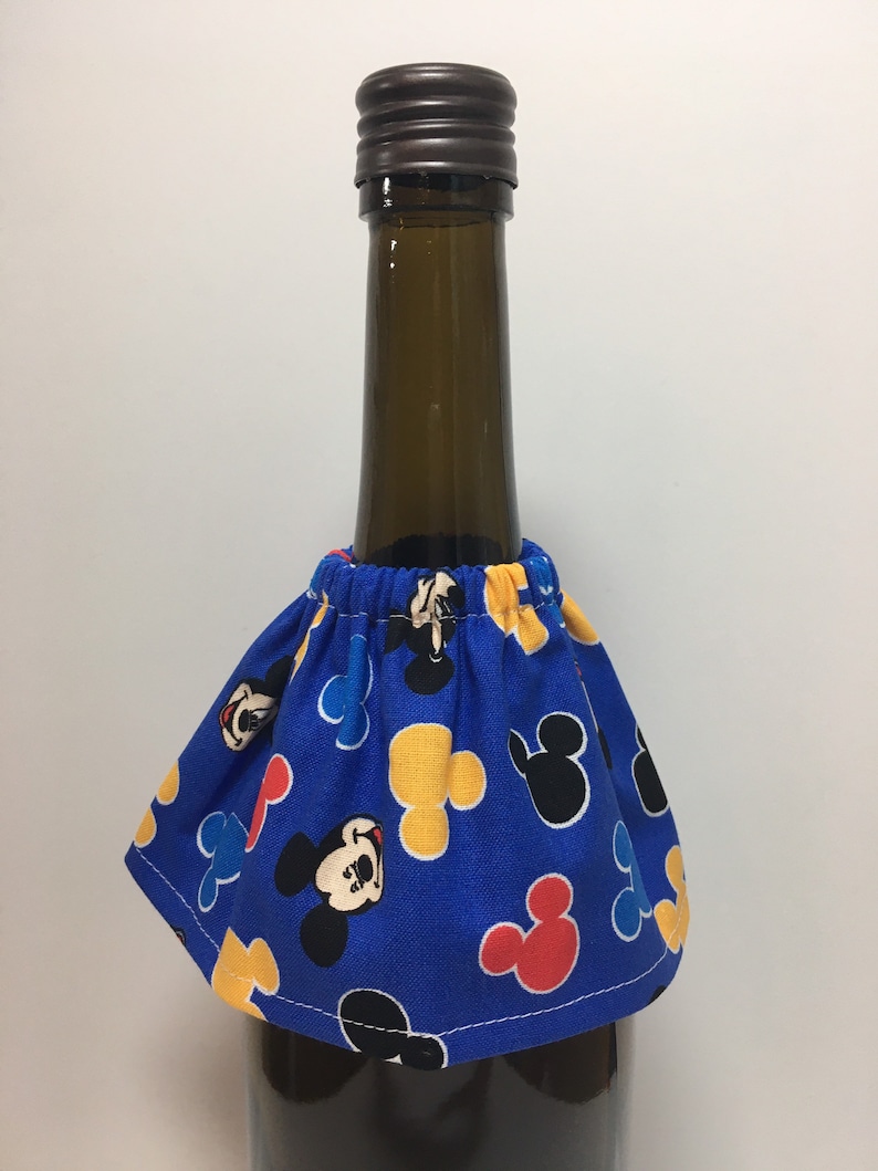 May include: A blue fabric wine bottle skirt featuring a pattern of Mickey Mouse faces and shapes in red, yellow, and black. The skirt is gathered at the top and fits around the neck of a wine bottle. The bottle has a brown cap.