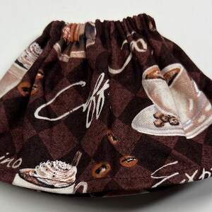 May include: A brown fabric coffee cup cosy with an elasticated band. The cosy features a repeating pattern of coffee cups, coffee beans, and the words "espresso" and "cappuccino" in white script. The background is a brown and dark brown chequered pattern.