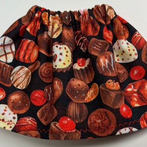 May include: A fabric item with an elasticated top, patterned with a repeating design of chocolates in brown, red, and white against a black backdrop. The item is shaped like a gathered skirt.