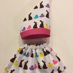 May include: A white fabric hat and skirt set with a repeating pattern of brown bunnies, and colorful chicks and eggs. The hat has a pink brim. The skirt has an elastic waistband.