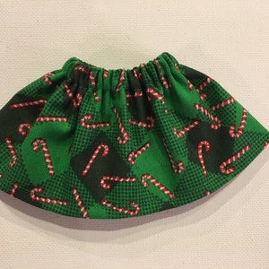 May include: A green fabric skirt featuring a repeating pattern of red and white candy canes. The skirt has an elasticated waistband and a flared shape, ideal for festive occasions. The fabric has a checkered pattern.