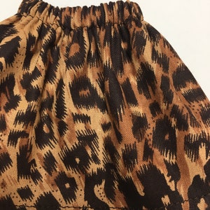 May include: A close-up of a skirt with an elastic waistband. The skirt features a brown and black leopard print pattern. The fabric is gathered at the waistband, creating a ruffled effect. The skirt is made of a lightweight material.