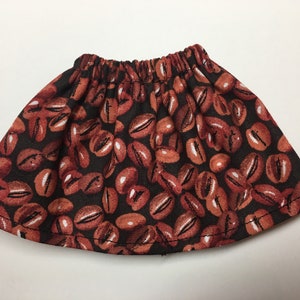May include: A small, gathered skirt with a coffee bean print. The fabric features a pattern of brown coffee beans with white highlights against a dark brown background. The skirt has an elastic waistband.