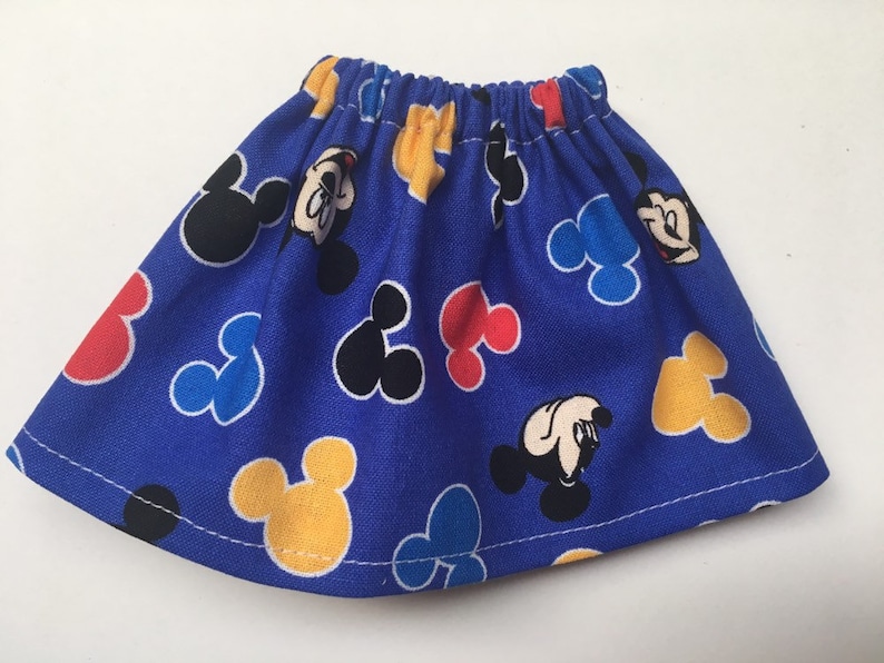May include: A blue skirt with an elastic waistband, featuring a pattern of Mickey Mouse heads in black, yellow, red, and blue. The skirt is made of a lightweight fabric and has a simple, gathered design.