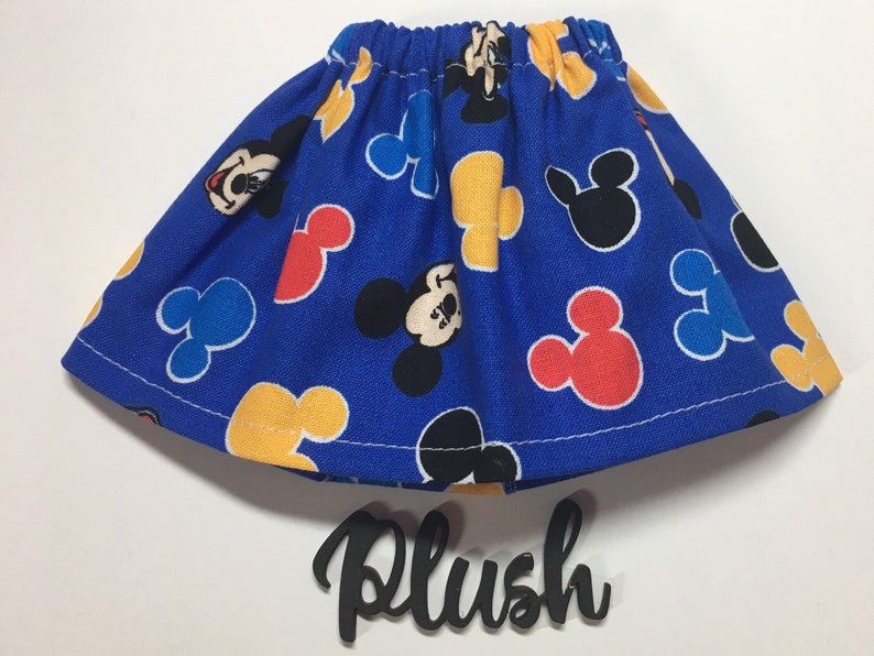 May include: A blue skirt with an elastic waistband, patterned with Mickey Mouse and Mickey Mouse-shaped designs in red, yellow, and black. The word "Plush" is written in a cursive font below the skirt.