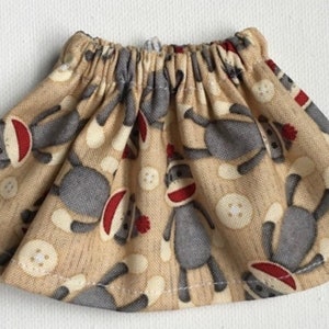 May include: A beige, gathered-waist skirt featuring a repeating pattern of gray sock monkey characters with red accents and white buttons. The skirt is made of a lightweight fabric, suitable for children's wear.