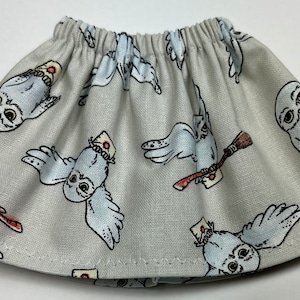 May include: A gray skirt with an elastic waistband, patterned with cartoon owls carrying letters and brooms. The owls are white with black outlines and are set against a light gray background. The skirt has a simple, gathered design.