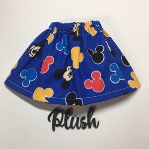 May include: A blue skirt with an elastic waistband, patterned with Mickey Mouse and Mickey Mouse head shapes in red, yellow, and black. The word "Plush" is written in black script below the skirt.
