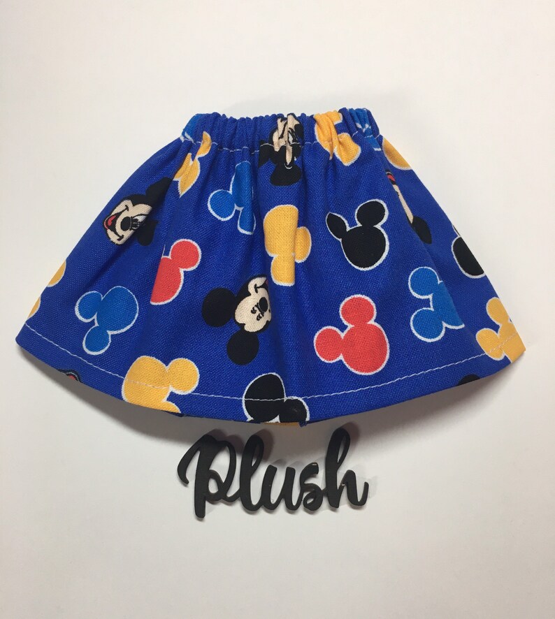 May include: A blue skirt with an elastic waistband, patterned with Mickey Mouse and Mickey Mouse head shapes in red, yellow, and black. The word "Plush" is written in black script below the skirt.