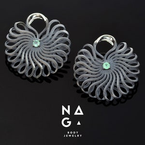 以下が含まれることがあります： 渦巻き状の花のようなデザインのシルバー製のイヤリングのペア。各イヤリングの中央には小さな緑色の宝石が埋め込まれています。