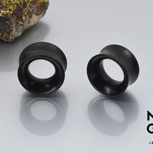 May include: Two black wooden ear gauges, also known as plugs or tunnels, are shown on a white surface. The gauges are round and have a slightly flared edge.