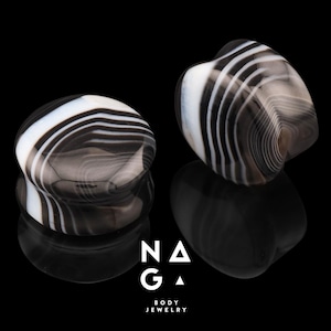 May include: Two black and white striped stone plugs for stretched ear piercings. One plug is round, the other is heart-shaped. The plugs are displayed on a reflective black surface. The logo "NAGA BODY JEWELRY" is at the bottom.