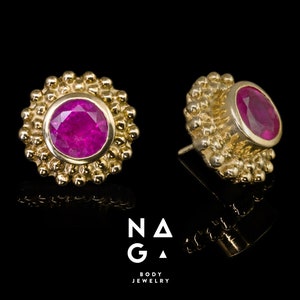 Könnte beinhalten: Zwei goldfarbene Ohrstecker mit einem leuchtend pinkfarbenen Edelstein in der Mitte. Die Ohrringe haben einen strukturierten, perlenbesetzten Rand. Die Ohrringe werden auf schwarzem Hintergrund präsentiert. Der Text "NAGA BODY JEWELRY" ist sichtbar.